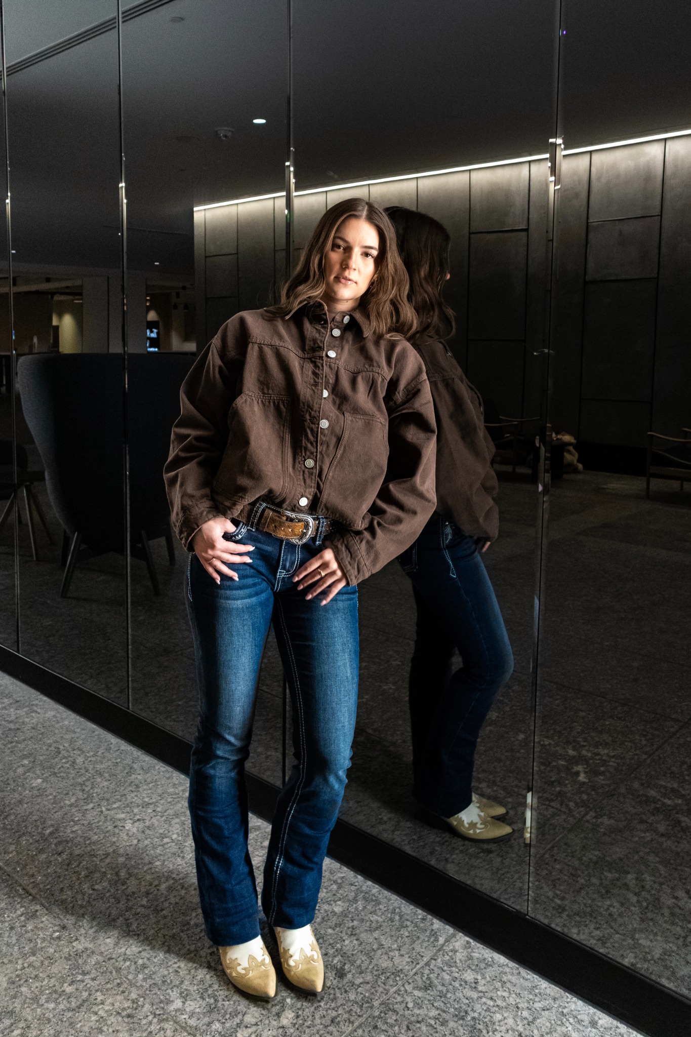 Keely wearing a brown jacket and jeans beside a mirrored wall